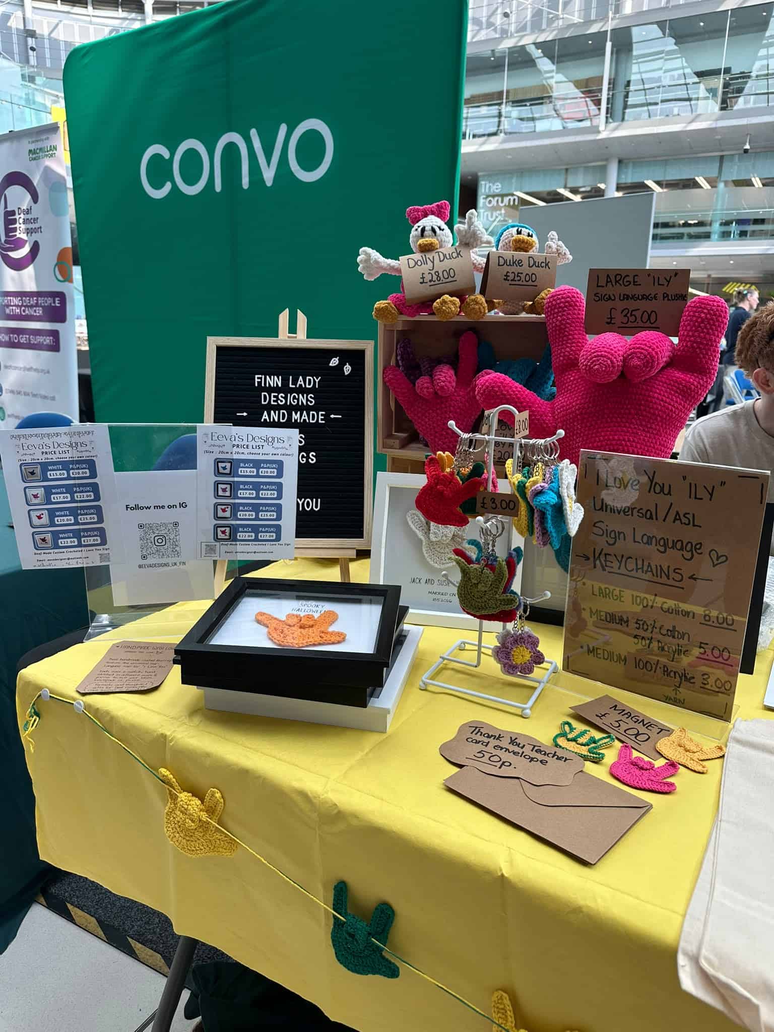 A craft stall displays crocheted "I Love You" BSL hand signs, keyrings, and pricing signs on a yellow tablecloth, with informational signs and QR codes.