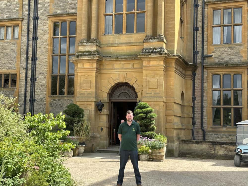 A person stands in front of a large stone building with tall windows and an arched doorway, surrounded by manicured plants.