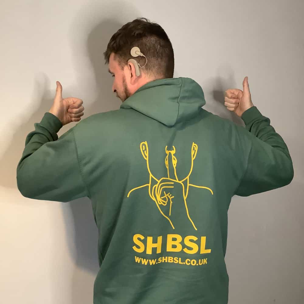 A man wearing a green hoodie with a yellow sign language design on the back and "SH BSL" website URL, giving thumbs up.