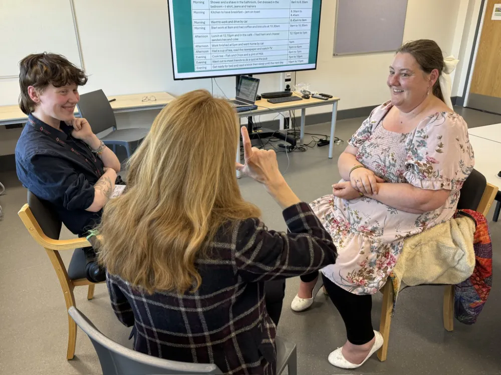Three people sit in a classroom in conversation, one using sign language. A large monitor displaying text is visible in the background.