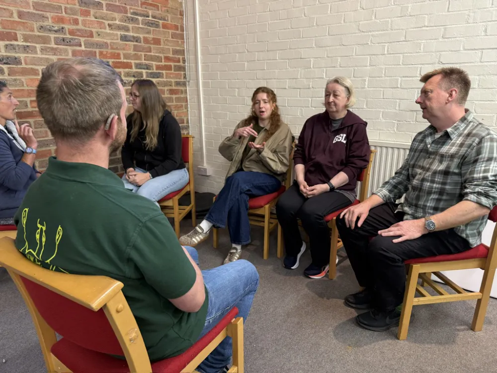 Six people sit in a circle on red chairs in a room with brick walls. One person is using sign language whilst others listen attentively.