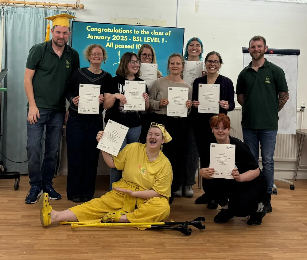 A group of ten people, some holding certificates, pose indoors in front of a sign congratulating the class of January 2025 for passing BSL Level 1. One person is seated on the floor with crutches.