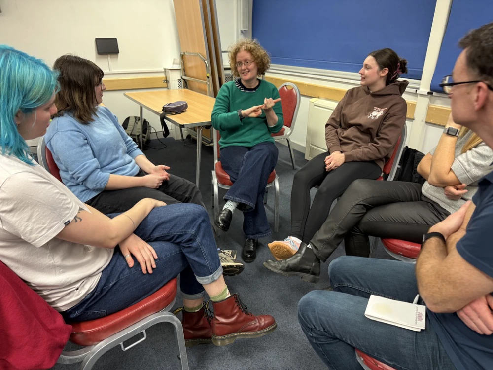 Six people sit in a circle in a classroom having a discussion. One person in a green jumper gestures whilst others listen attentively. A table and blueboard are in the background.