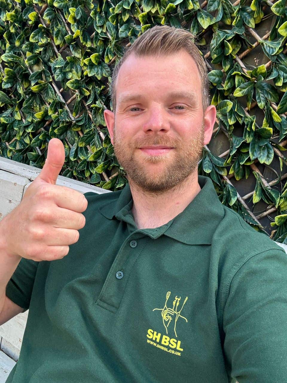 A man with a beard gives a thumbs up, wearing a green polo shirt with a tree logo, standing against a leafy wall.