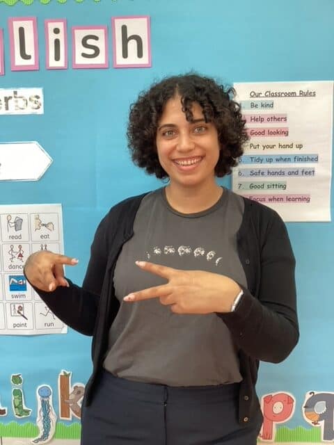 A person with curly hair smiles and gesturing with both hands. They're standing in front of a classroom bulletin board displaying educational materials.