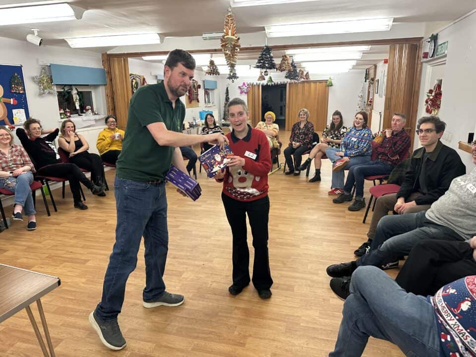 A man hands a gift to a smiling woman in a festive sweater, while seated people watch in a decorated room adorned with Xmas lights.