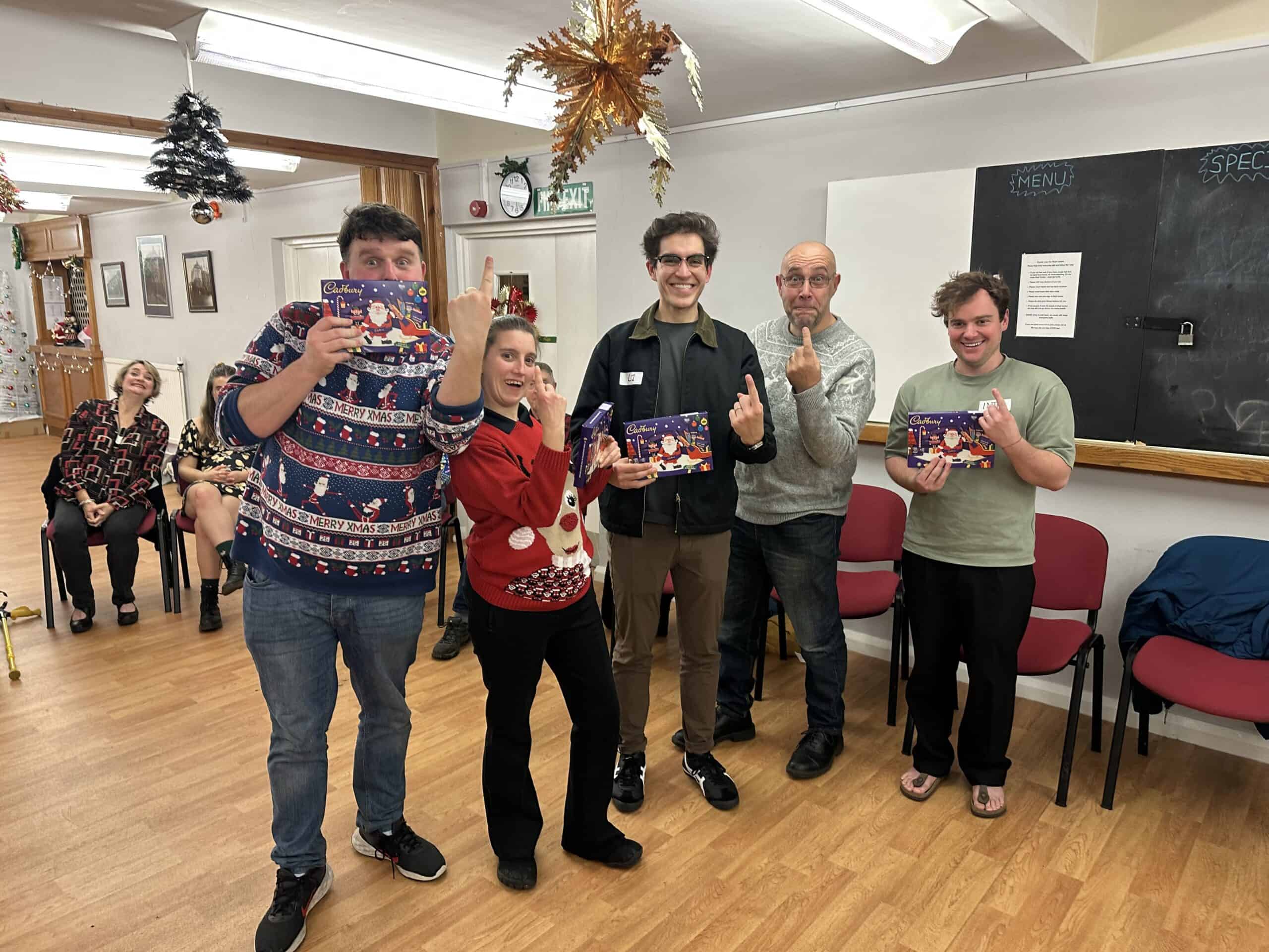 Five people standing indoors, holding chocolate gifts and posing playfully. Holiday decorations are visible, including a Christmas tree and ornaments. Three people are seated in the background.