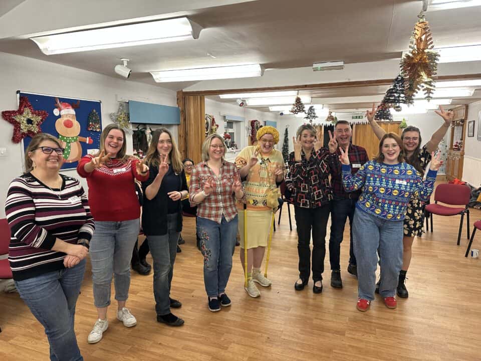 A group of people posing indoors, some wearing festive sweaters for Xmas. Holiday decorations hang from the ceiling and walls, creating a cheerful atmosphere.