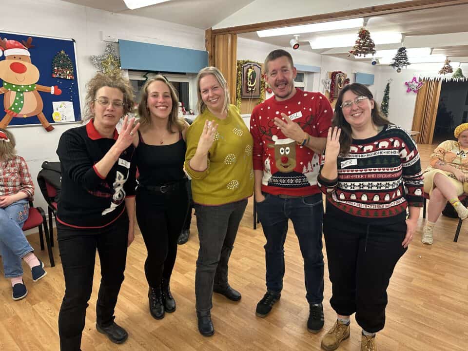 Five people in festive sweaters stand together indoors, smiling and holding up four fingers in what looks like a BSL sign. Christmas decorations are visible in the background, adding to the joyful atmosphere.