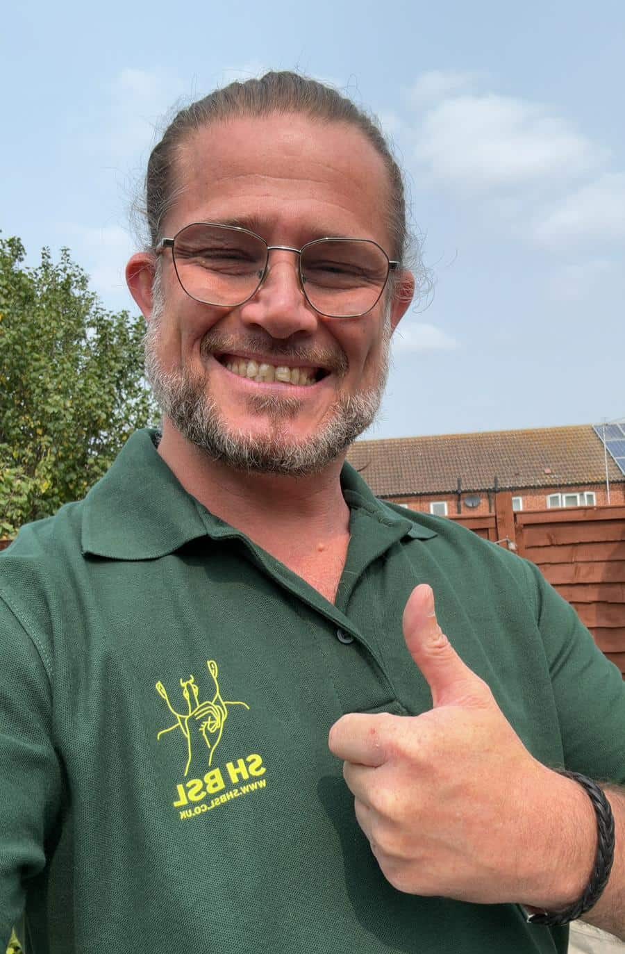 Man with glasses smiles and gives thumbs up. Wearing a green polo shirt with a yellow logo. Background includes trees and buildings.