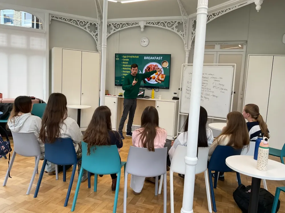 A teacher stands at the front of a classroom, speaking to a group of pupils seated in chairs, with a screen displaying a breakfast menu behind him.