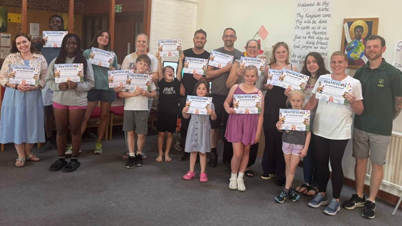 A group of people, both adults and children, stand indoors holding certificates and smiling at the camera.