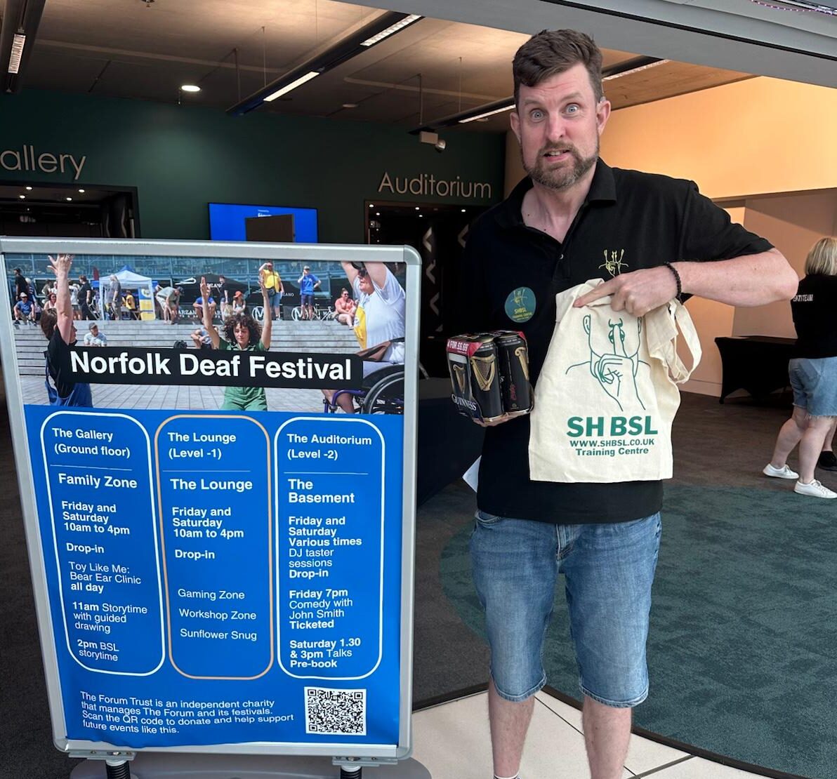 A man stands indoors next to a Norfolk Deaf Festival sign, holding a tote bag and a packaged item, with an event venue visible in the background.