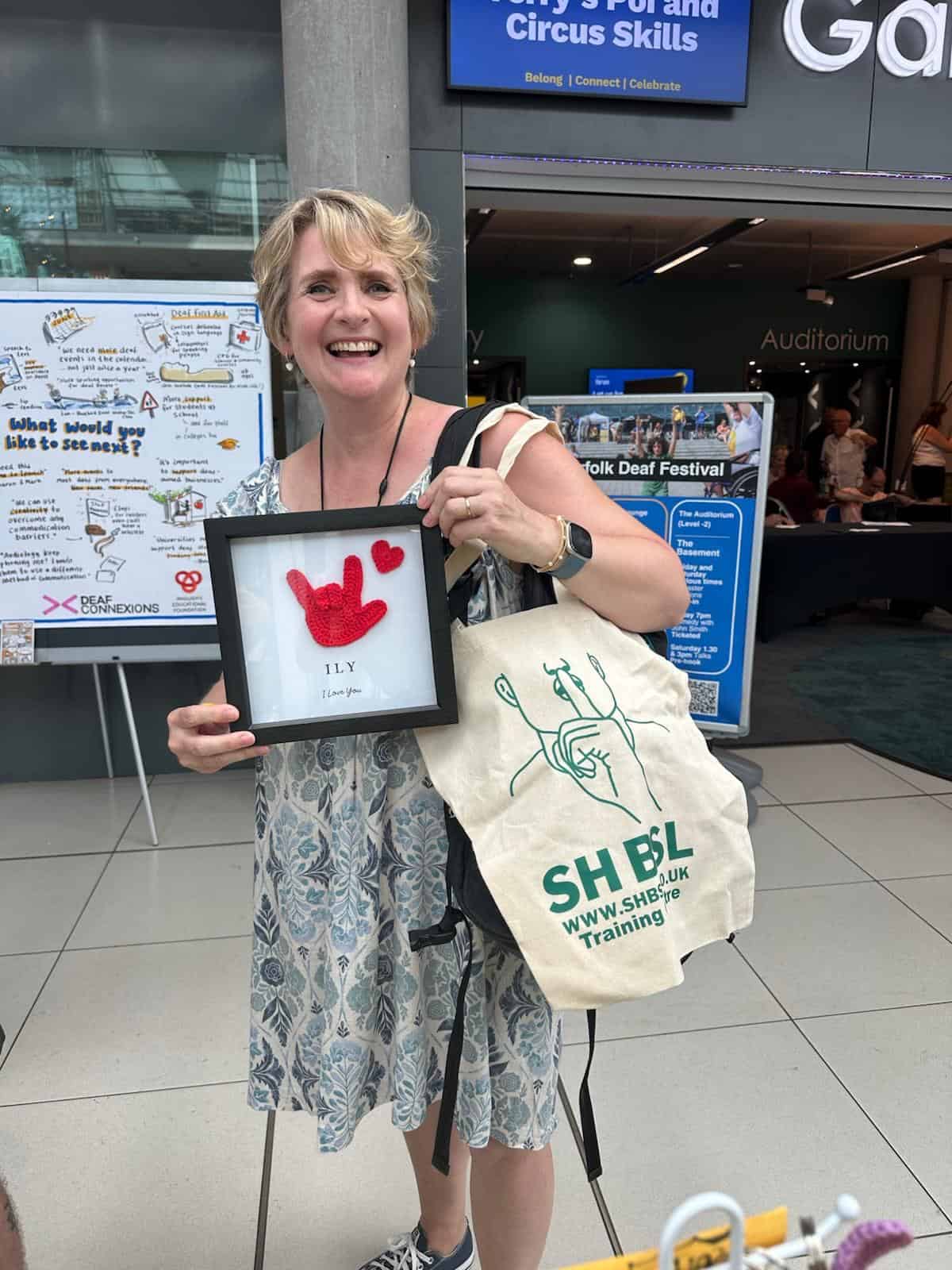 A woman smiling and holding a framed picture of the American Sign Language "I Love You" sign, standing indoors with an SHBSL tote bag.