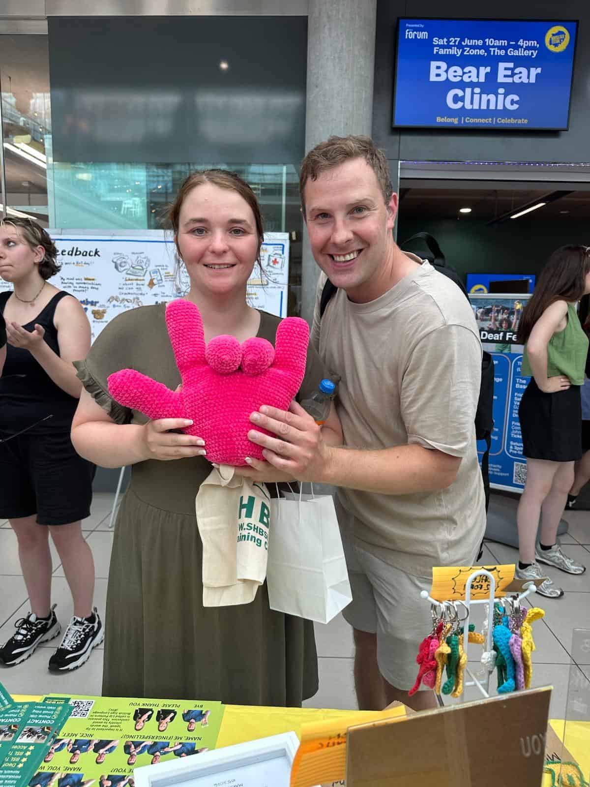 Two people smiling and holding a pink soft toy at an indoor event table, with informational posters and other attendees visible in the background.