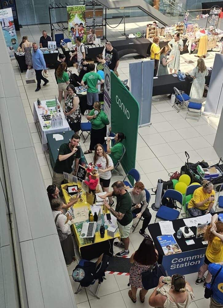 A busy indoor event with multiple stands and tables, people interacting, and informational displays set up in a spacious, modern venue with glass walls.