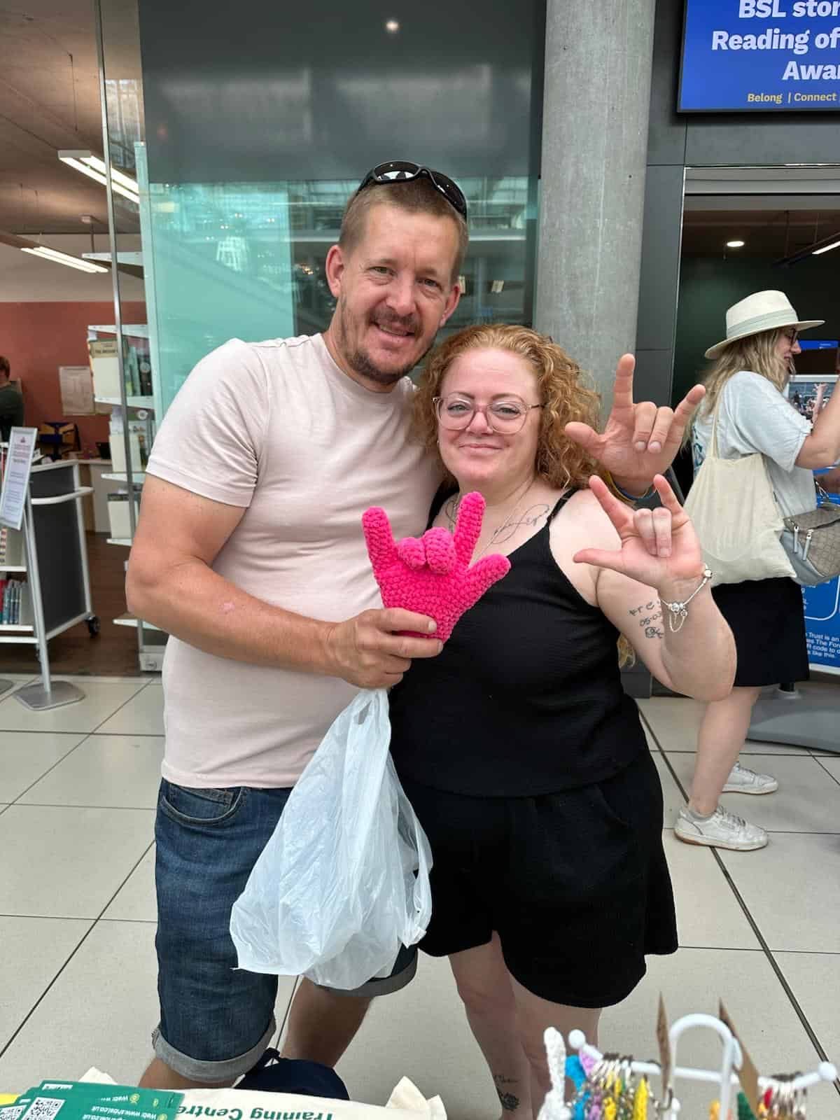 Two people stand indoors, smiling at the camera. One holds a pink knitted hand, both making the "I love you" sign in sign language. Other people are visible in the background.