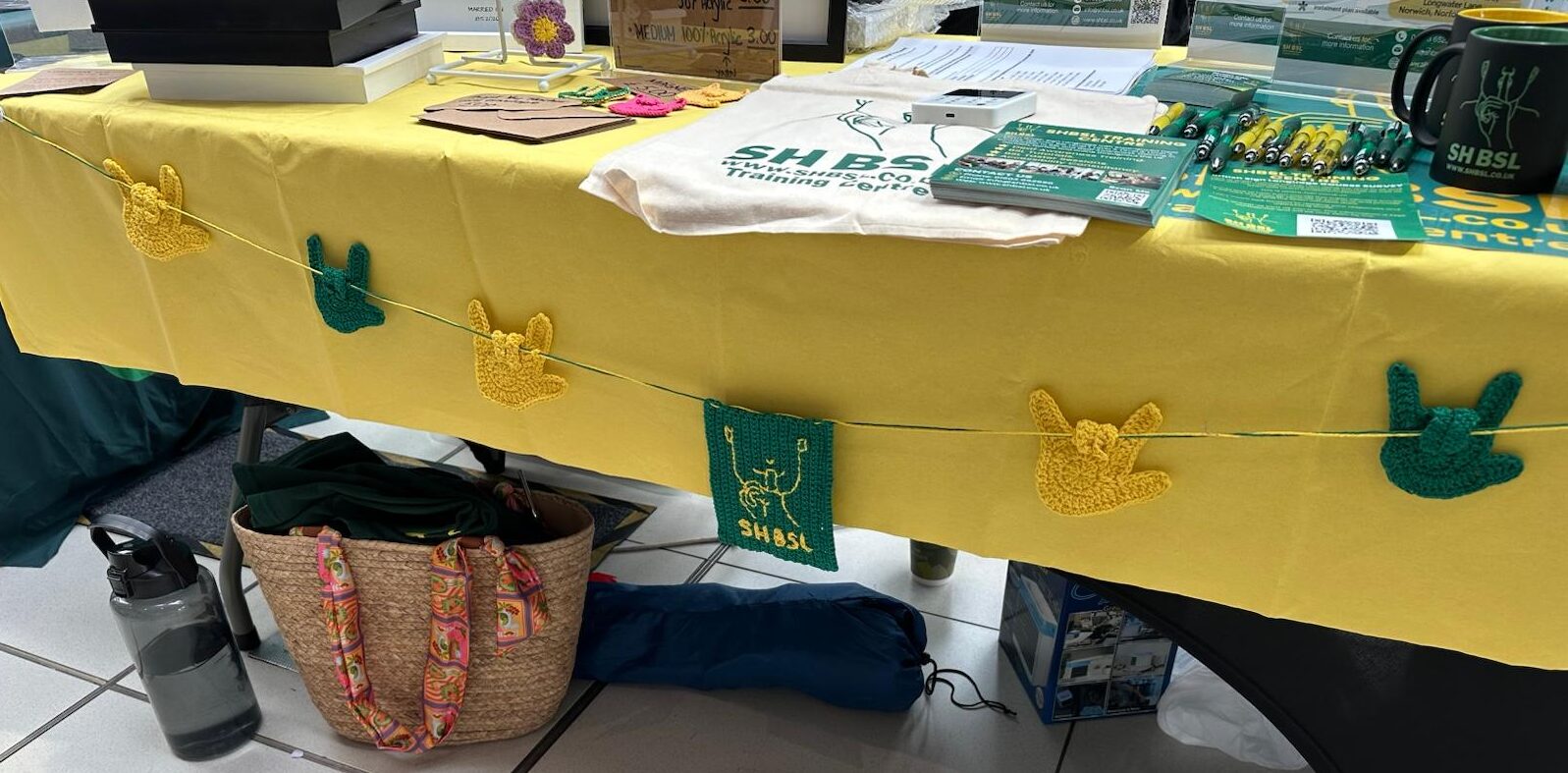 A yellow tablecloth with green and yellow crochet decorations covers a display table. Under the table are a woven bag, water bottle, and other items.