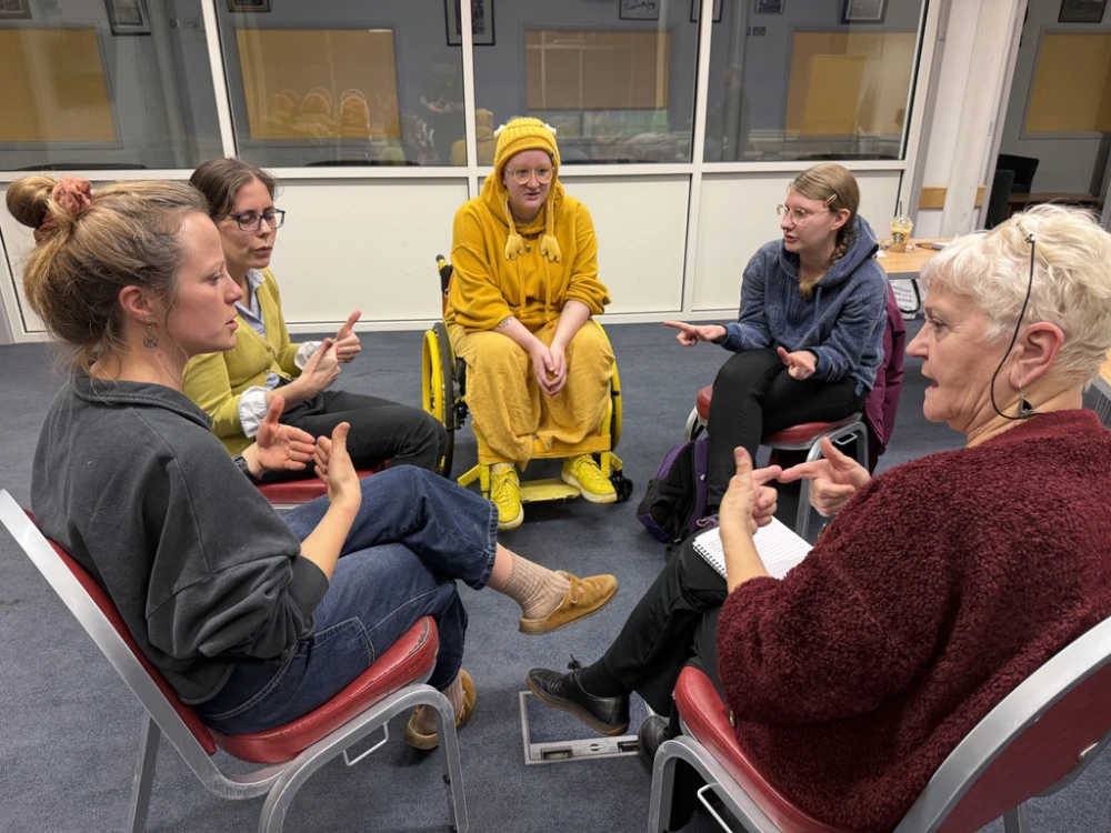 Six people sit in a circle indoors, engaged in conversation, with one person in a yellow outfit and wheelchair; some use hand gestures whilst one writes in a notebook.