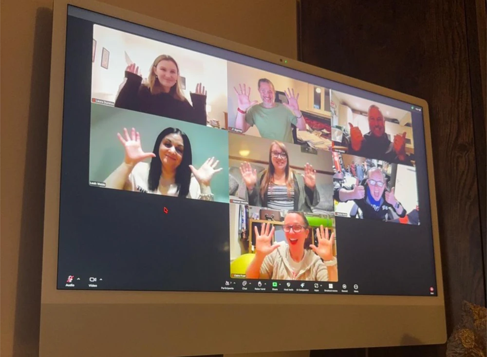Six people participate in a video call, each smiling and raising both hands towards the camera in individual video boxes on a screen.