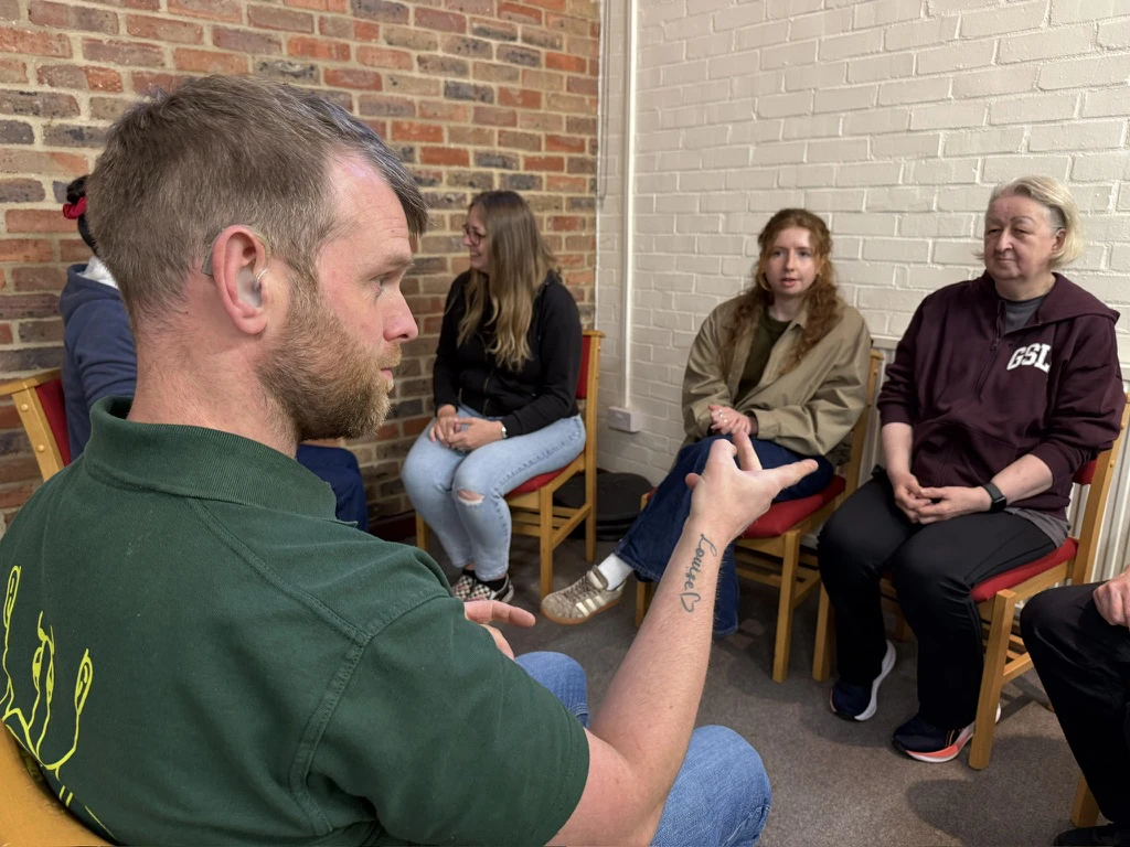 A man with a cochlear implant signs to a seated group of women in a room with brick and white walls.