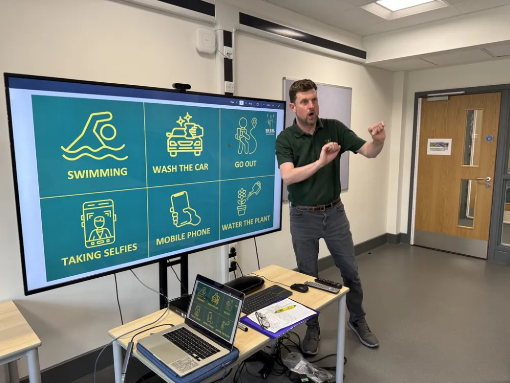 A man stands by a presentation screen displaying icons and text for activities: swimming, washing the car, going out, taking selfies, using a mobile phone, and watering plants.