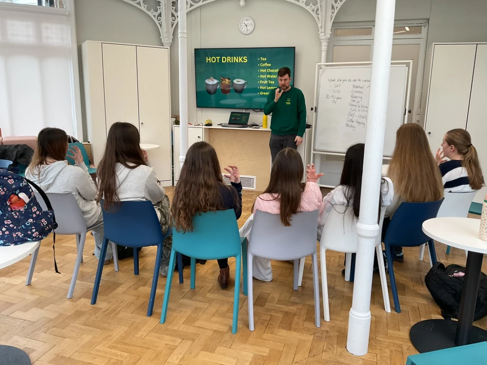 A group of students sits on chairs facing a man standing near a screen displaying a hot drinks menu in a classroom setting.