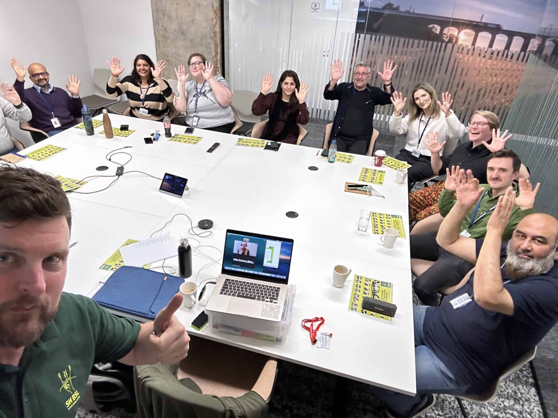 A group of people sitting around a conference table, waving at the camera. A laptop is open in the center of the table, connected to a video call.