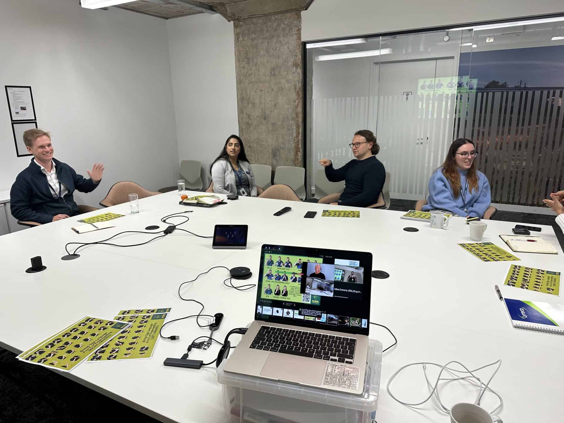 A group of people sit around a conference table with laptops, notes, and drinks, participating in a video call meeting.