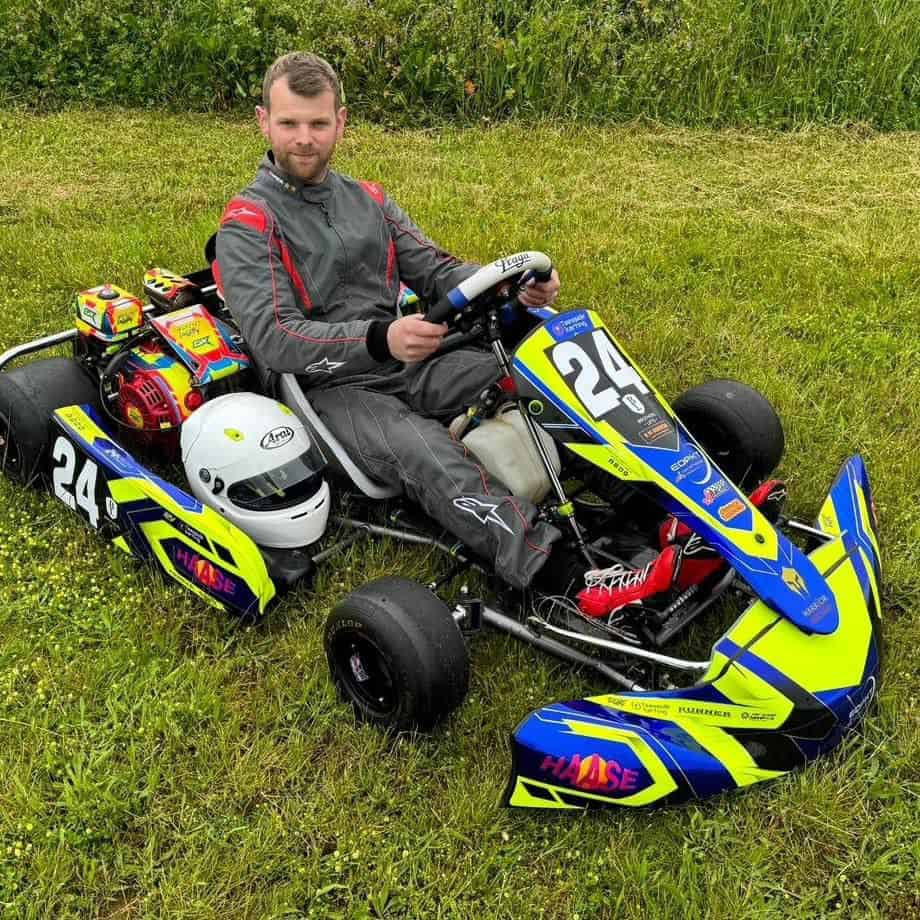 A person in a racing suit sits in a go-kart on the grass in Essex, surrounded by helmets and gear, looking ready to tutor anyone eager to learn the art of speed.