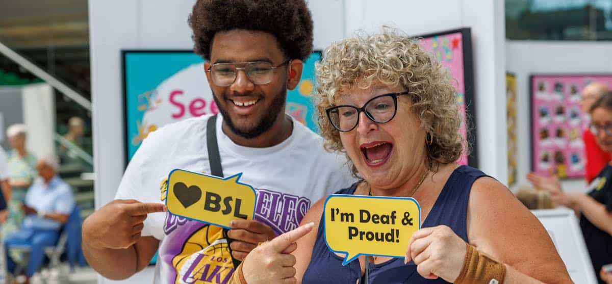 Two people smiling and holding signs; one sign reads "♥ BSL" and the other reads "I'm Deaf & Proud!" in a brightly lit indoor setting.