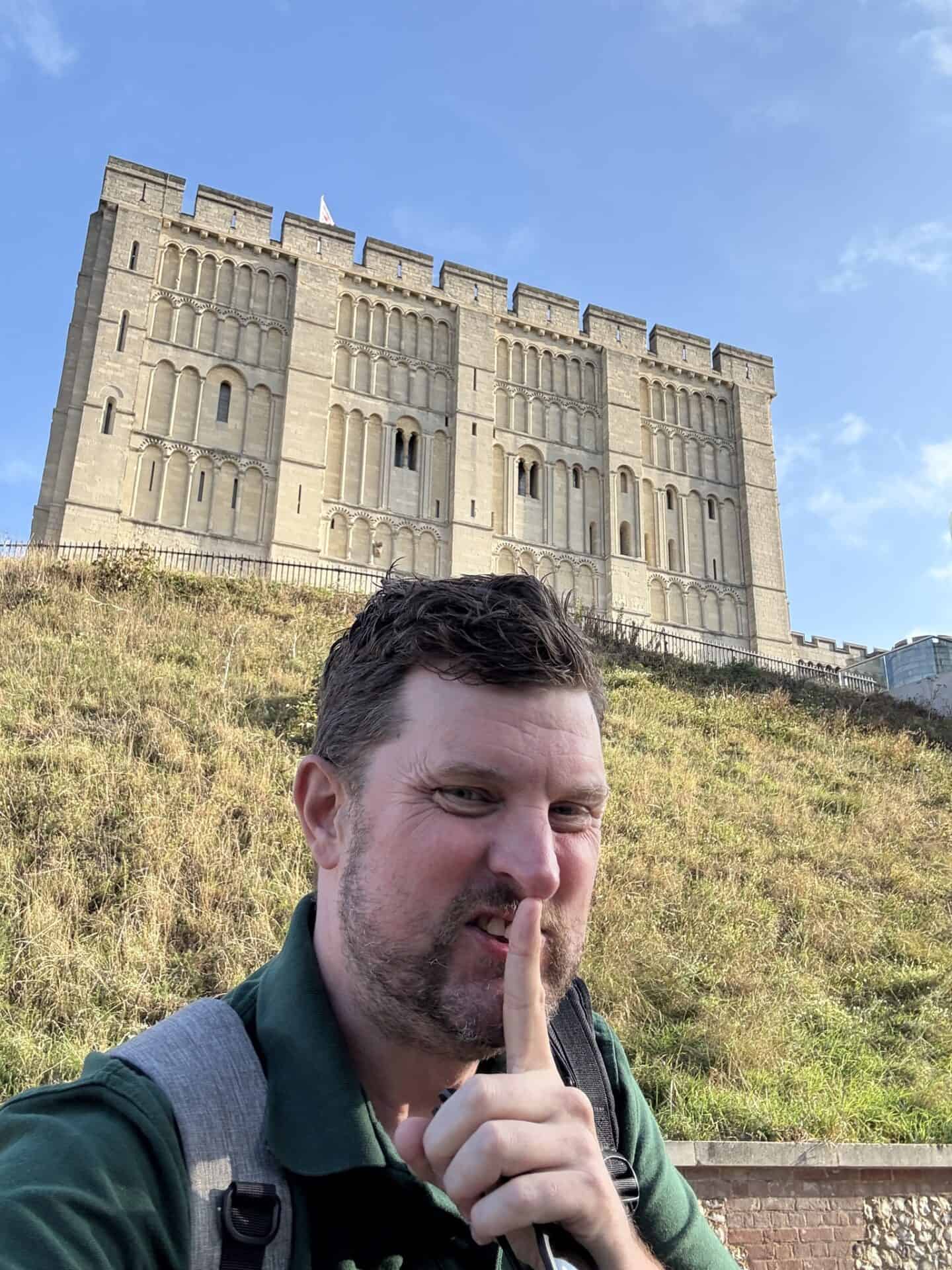 A man with a backpack holds a finger to his lips in a shushing gesture, standing in front of a large stone castle on a grassy hill under a blue sky.