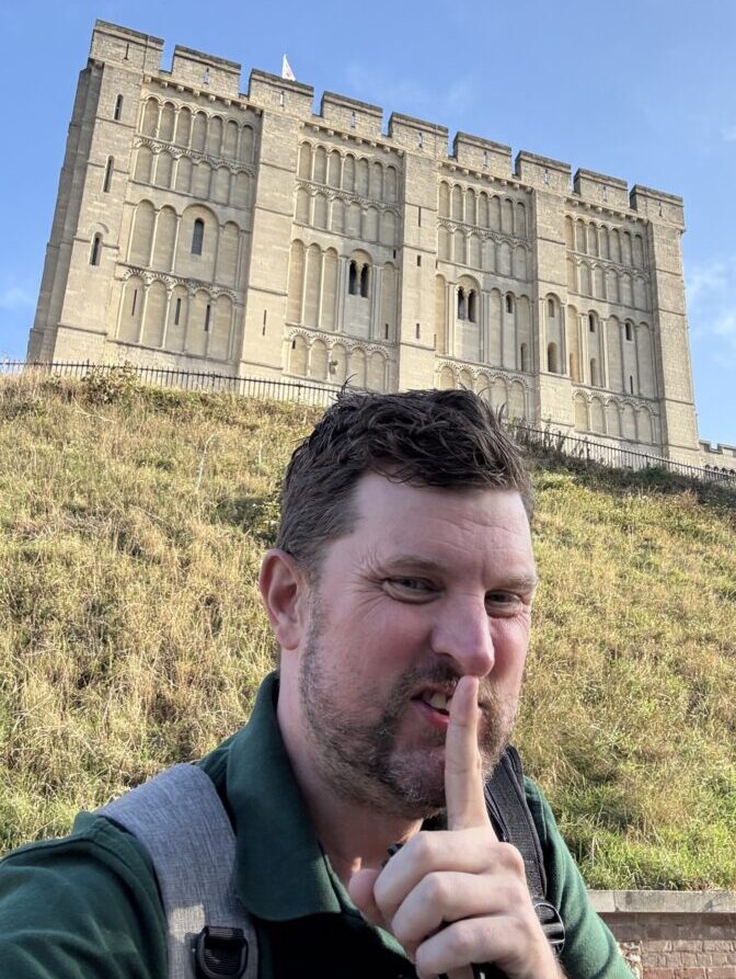 A man with a backpack holds a finger to his lips in a shushing gesture, standing in front of a large stone castle on a grassy hill under a blue sky.
